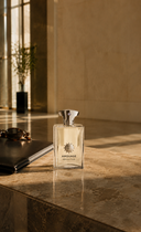 A clear perfume bottle with a silver cap beside a notebook and sunglasses on a sunlit stone surface.