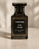 A 50 ml Tom Ford Oud Wood Eau de Parfum bottle, made of dark amber-brown glass with a black cap and a black label featuring white text, set against a warm beige background with soft leaf shadows.