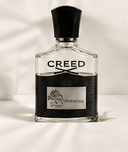 A bottle of Creed Aventus fragrance with a black cap, clear glass body, and a black-and-silver label, photographed against a softly lit white background.