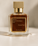 A bottle of Maison Francis Kurkdjian Grand Soir Eau de Parfum, featuring a clear glass bottle with amber liquid, a gold cap, and a brown label with gold lettering, set against a warm beige background.