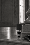 A bottle of black ink with a dark wood-textured cap and a clear glass base, standing on a stone ledge in a monochrome interior setting.