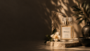A clear glass perfume bottle labeled BELLA by Scent of the Sea stands on a stone slab in a styled setting with warm sunlight, leaf shadows, a white flower, and dark green leaves.