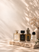 Four perfume bottles arranged on a stone slab against a warm beige wall with leaf shadows. The bottles include Bella, Fabrice Black Oak, X, and Sal d'Afrique, with dried flowers in a ceramic vase beside them.